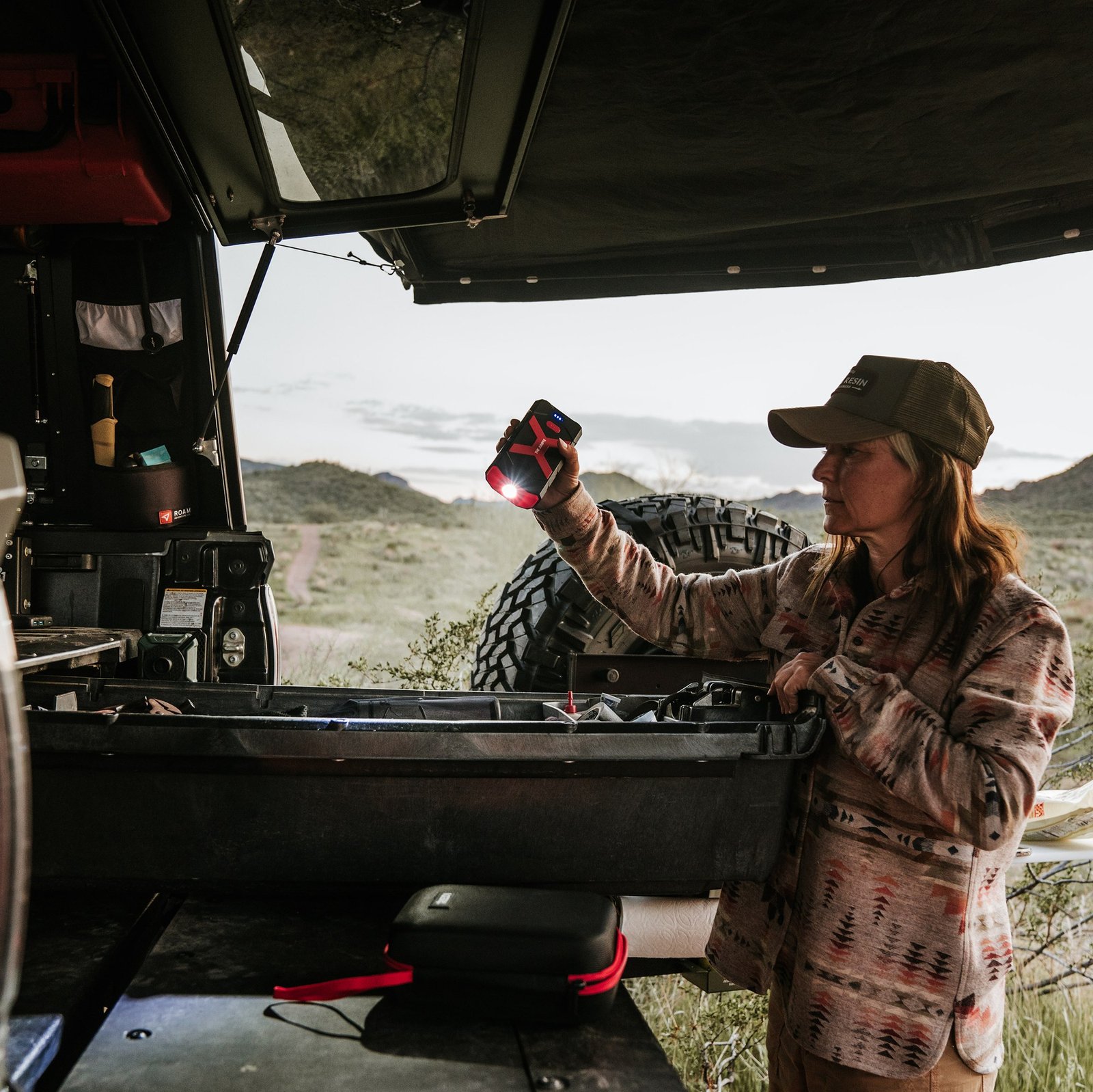 Person using the Redi Jumpie flashlight near an open vehicle cargo area with the hard-shell case on a tailgate