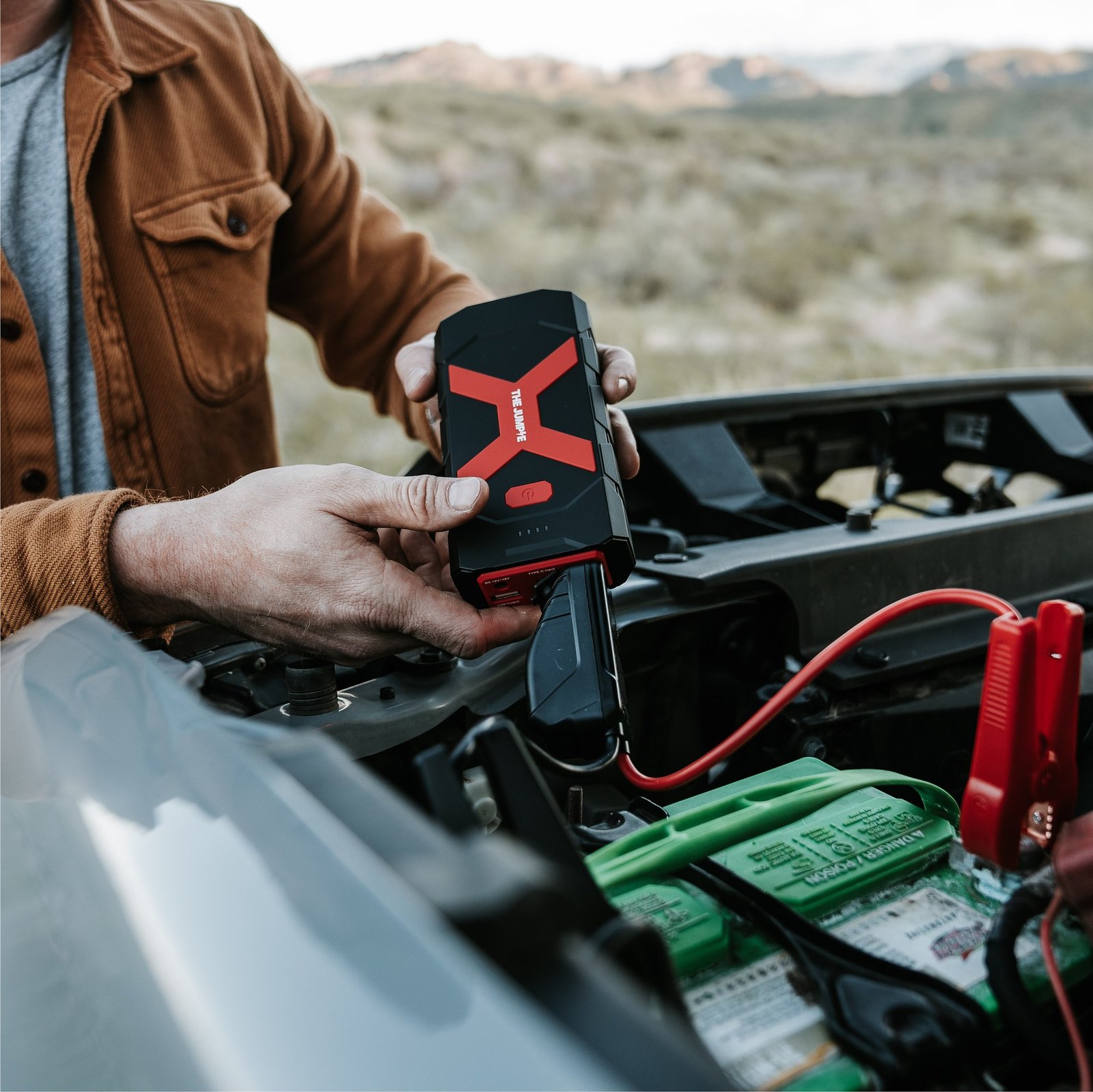 Person holding a Redi Jumpie beside an open hood with jumper clamps on a vehicle battery
